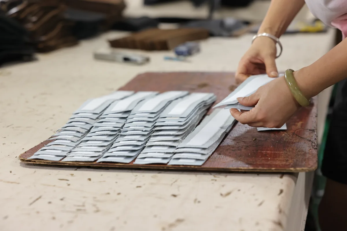 Hands sorting rows of leather bag components on a factory workbench