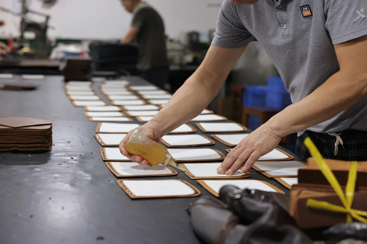 Hands applying adhesive to rows of leather panels with white backing at a factory table