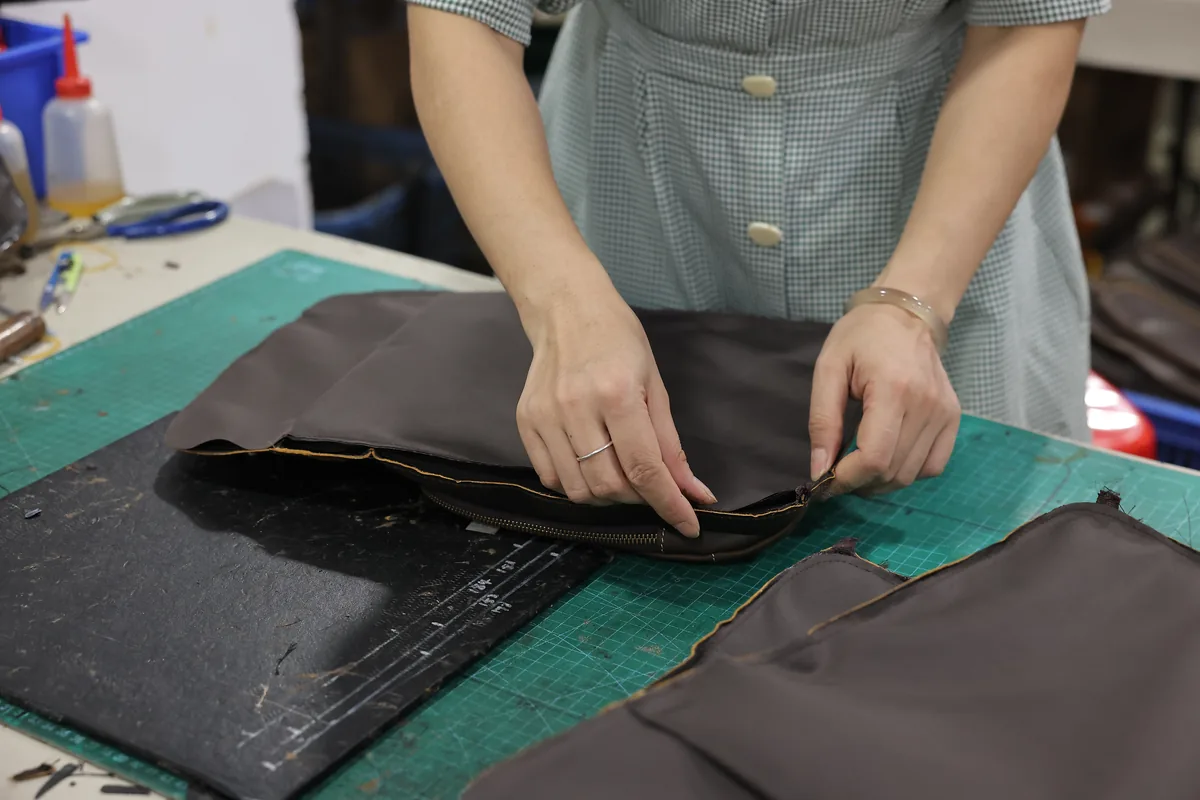 Hands arranging a zipper along a leather bag panel at a factory workbench