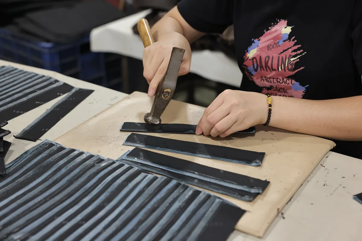 Hands using a hammer to work on leather components at a factory workbench