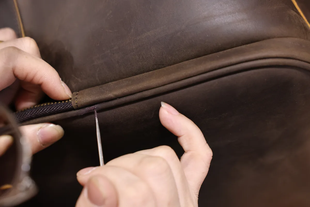 Hands checking a zipper seam on a dark brown leather bag